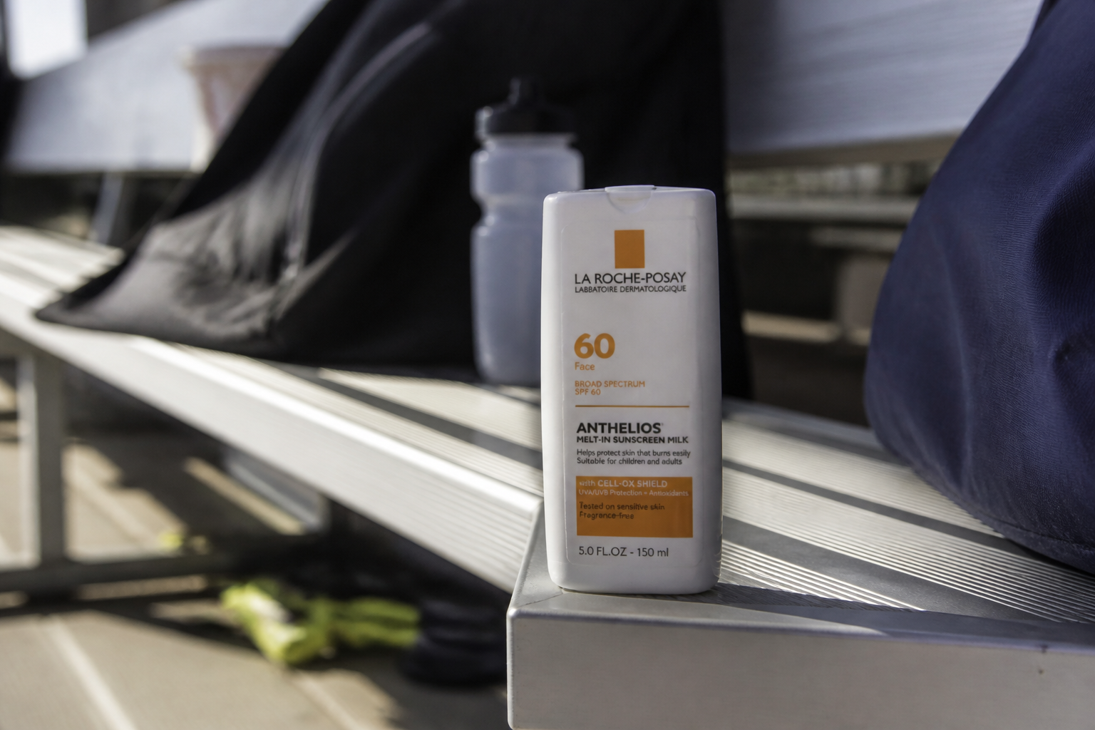Botella de protector solar SPF 50 junto a gorra blanca, gafas de sol y bálsamo labial en un banquillo de pista al aire libre