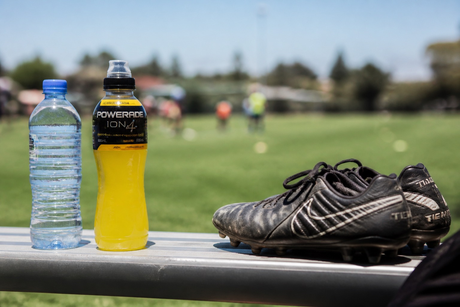Botella de agua e isotónica junto a botas de fútbol en un banquillo de campo al sol